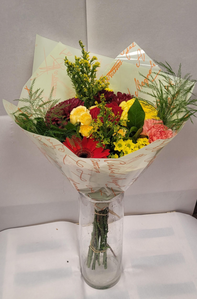 Sunset Garden Bouquet - A lively bouquet showcasing a rich mix of warm, sunset-inspired colors. Deep red and bright yellow blooms take center stage, complemented by soft pink flowers, fresh greenery, and delicate filler stems. Wrapped in a light cream paper with decorative script, the arrangement sits in a clear glass vase, creating a cheerful and elegant display perfect for celebrations, gratitude, or brightening someone’s day.