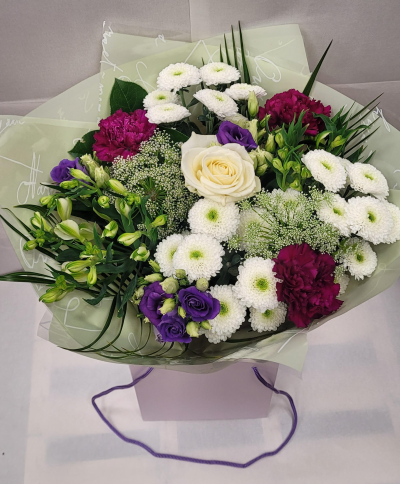 Lavender Bliss Bouquet - A graceful bouquet featuring a soothing palette of white, cream, and rich purple. At its center rests a creamy white rose surrounded by fluffy white chrysanthemums, deep magenta carnations, and elegant purple lisianthus blooms. Soft green buds and feathery filler flowers add texture and freshness, while long, slender foliage enhances the arrangement’s natural flow. Presented in pale green wrapping and placed in a lilac gift box, this bouquet evokes charm, calm, and refined beauty—perfect for thoughtful gestures, sympathy, or celebrations of elegance.