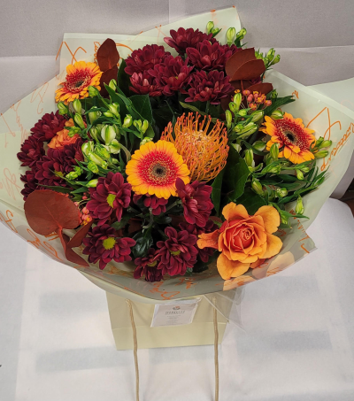 Autumn Glow Bouquet - A vibrant arrangement inspired by the warm hues of fall. Rich burgundy chrysanthemums pair beautifully with bright orange gerbera daisies and a striking orange rose, creating a bold and cheerful blend. Unique pincushion protea adds texture and flair, while fresh greenery and rustic brown leaves enhance the seasonal feel. Wrapped in soft cream paper and presented in a matching gift bag, this bouquet radiates coziness and celebration—perfect for autumn occasions, heartfelt thanks, or adding a burst of warmth to any space.