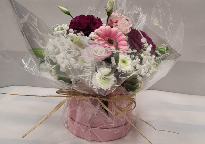 Sweet Serenity Hatbox - A delicate and elegant floral arrangement presented in a charming pink hatbox. Soft pastel blooms, including pink gerbera daisies, blush roses, and white chrysanthemums, are complemented by rich plum carnations and dainty sprays of baby's breath. Wrapped in clear decorative film and tied with a natural raffia bow, this arrangement exudes a sense of calm and tenderness. Perfect for expressing love, gratitude, or gentle affection, Sweet Serenity Hatbox makes a timeless and graceful gift for any occasion.