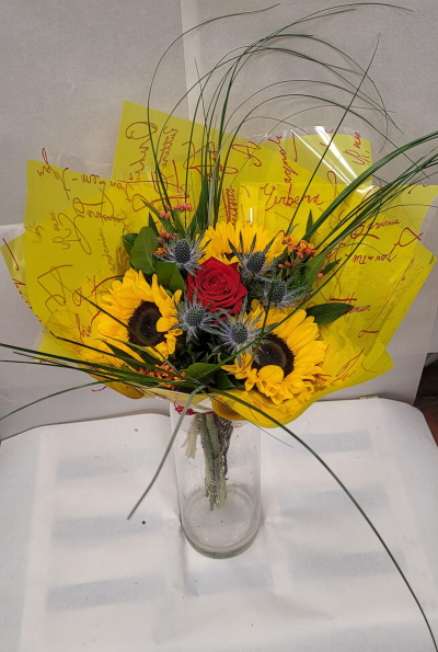 Radiance - A radiant bouquet bursting with warmth and contrast. Golden sunflowers take center stage, their bright petals glowing against deep green foliage. A single red rose adds a touch of romance and intensity, while blue thistles lend unique texture and charm. Wisps of decorative grass create graceful movement, all wrapped in vibrant yellow paper with red script detailing. This striking arrangement captures the joy of sunshine and the elegance of love—perfect for celebrations, congratulations, or heartfelt gestures.